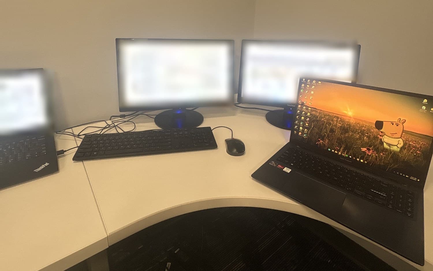 A corner office desk with two monitors and a docked ThinkPad, their screens out of focus, a keyboard and mouse in front, and a personal laptop open at the right.